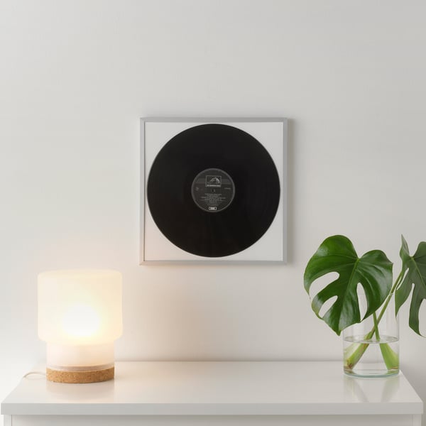 A framed black vinyl record hung on a light grey wall above a white shelf. The shelf holds a small lamp and a glass vase with green leaves.