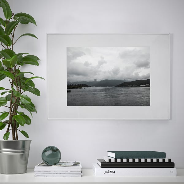 White LOMVIKEN picture frame with black and white maritime image, placed above books and a decorative object. Green plant to the left.