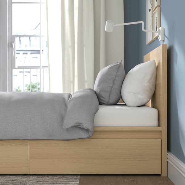 Bed by window, grey comforter, pillows, headboard with wall lamp.