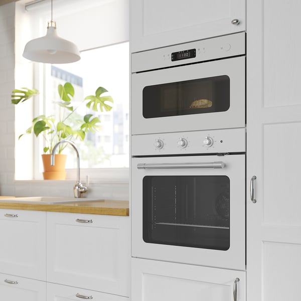 Modern kitchen with white built-in double oven, digital display on top oven, minimalist design; potted plant by a window.
