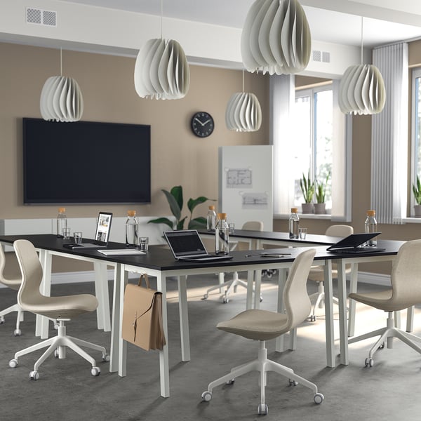 Modern office conference room with MITTZON tables, beige chairs, white pendant lights, black tv, and clock. Tables large, with laptops and cups.