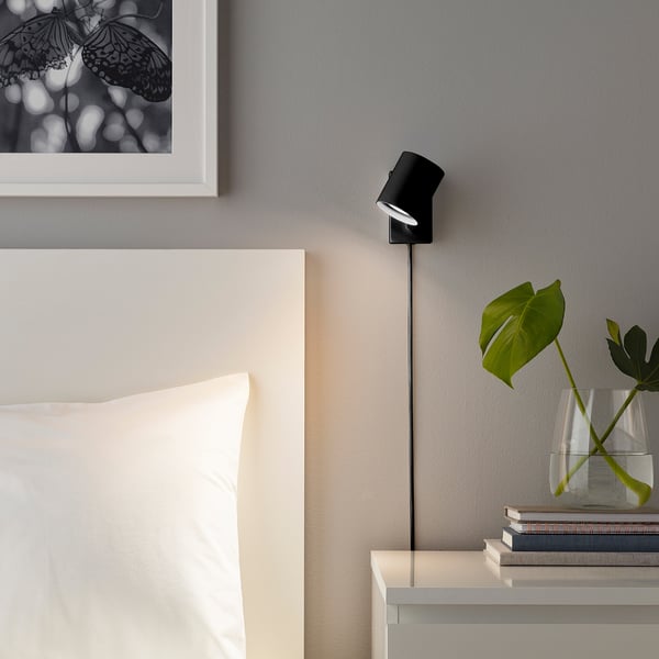 Wall-mounted adjustable light, NYMÅNE, beside bed, emits warm light, near plant and picture frame.
