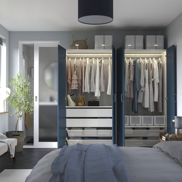 Modern bedroom with open PAX wardrobes displaying clothes, drawers, and storage boxes. Interior lighting highlights organised apparel and accessories.
