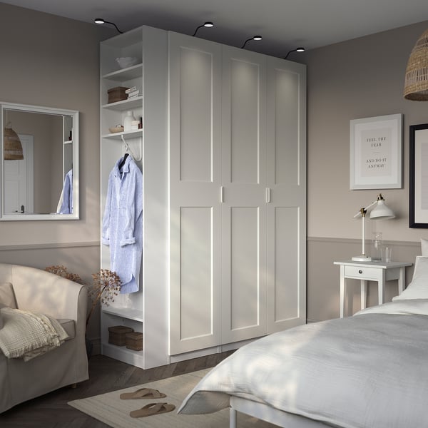 Modern bedroom with PAX wardrobe showing clothes, bed, armchair, and lamp.