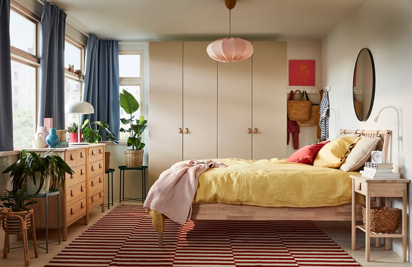 Bright, cosy bedroom with yellow bedding, wooden dresser, striped rug, hanging plants, and a large mirror.