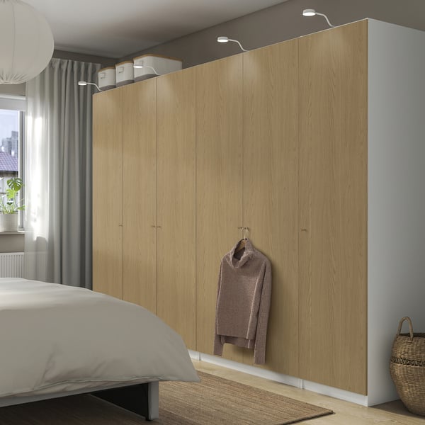 A large, light wood-colored PAX wardrobe against a bedroom wall. It has several doors and a hanging jacket, with built-in lighting.
