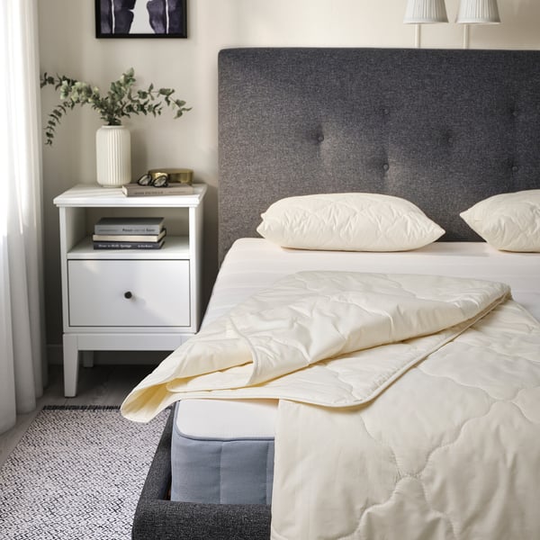 Minimalist bedroom with RÅDHUSVIN duvet on bed. Duvet is white, oversized, and made of cotton and wool. Bedside table nearby with books and vase.