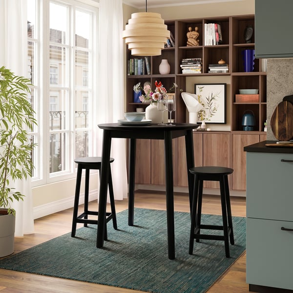 Modern black RÄVSTEN counter table and stools by window, with large rug, houseplant, and wooden shelves filled with books and decor.