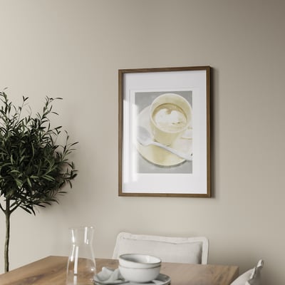 A coffee cup hangs on a wall above a wooden table with a white chair. A plant and carafe are on the table. The frame holds a picture up to 50x70 cm.