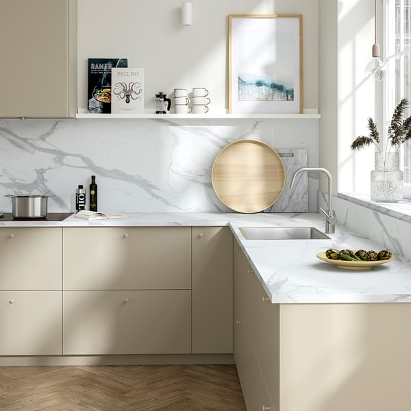 Modern kitchen with white marble countertops, beige cabinets, and a large round wooden tray. Artwork and greenery decorate the marble backsplash.