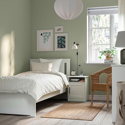 Cosy bedroom with SONGESAND bed, sage walls, crisp linens, rug, bedside table, lamp, plant, wicker chair, art, and large window.