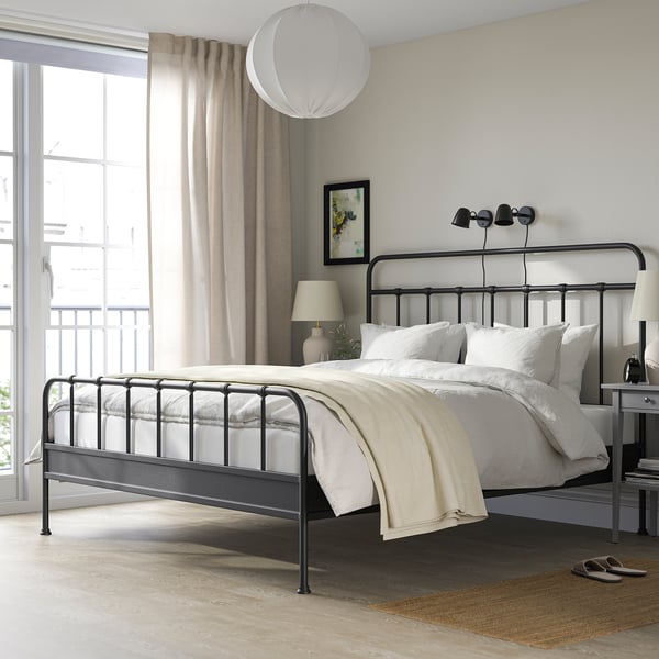 STJÄRNÖ black metal bed with curved headboard, white linens, high footboard. Minimalist room with large window, small rug.
