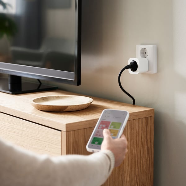 A person holding a smartphone with a home automation app, controlling a white smart plug connected to a wooden cabinet, with a bowl and a tv on top.