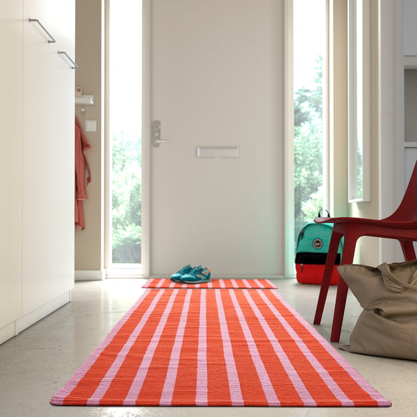 Bright orange and pink striped VÄGSKYLT rug, larger than the door mat beside it, welcomes at the entrance.