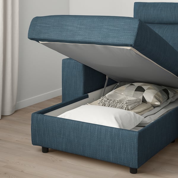 Blue storage sofa bed, open compartment showing folded sheets, white wall behind.