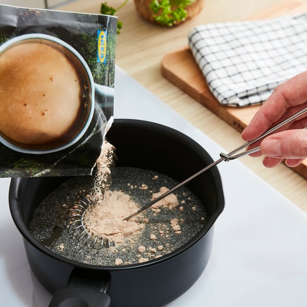 Personen häller ljusbrunt pulver i svart skål med metallsked, träyta, trasa, växt.