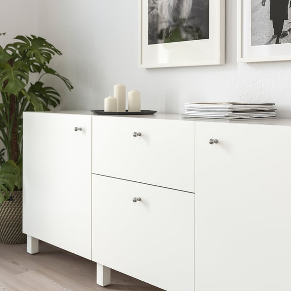 Modernt vitt skåp med metallknoppar, som rymmer växter, ljus och böcker, placerat under inramad väggkonst.