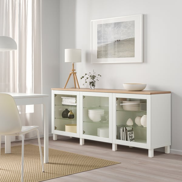 Vitt ikea BESTÅ-sideboard med glasdörrar, som visar fat och vaser, tillsammans med en lampa, en växt och en strandbild i en ljus matsal.