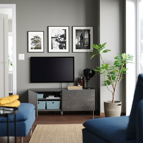 Modernt vardagsrum med grå BESTÅ tv-bänk, blå soffa och stor växt. Tv och inramade foton på väggen.