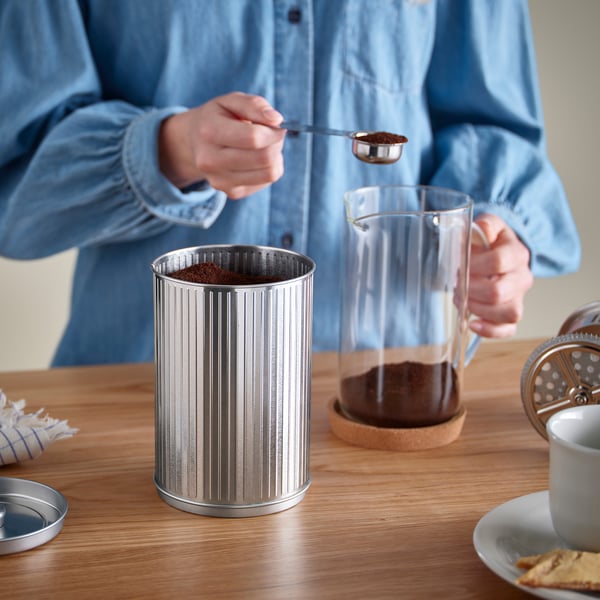 En person skopar upp kaffesump i en metallbehållare på ett bord, med en fransk glaspress och en mugg i närheten.
