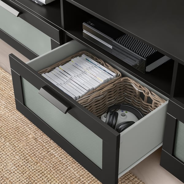 Svart tv-möbel med öppen bred låda som avslöjar flätad korg med videospelsfodral och kontroller, vilket visar upp organiserad förvaring.