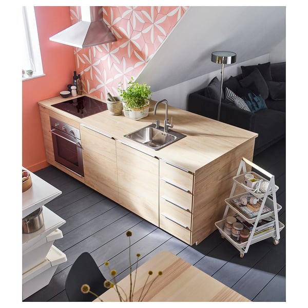 Modernt kök med bänkskiva i ljust trä, rostfri diskbänk, svarta vitvaror, rosa väggar, geometrisk stänkskydd, växt.