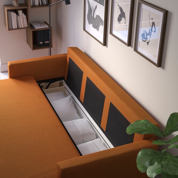 Modern orange bäddsoffa med öppen förvaring, som visar kuddar, mot vägg med konst och växt.