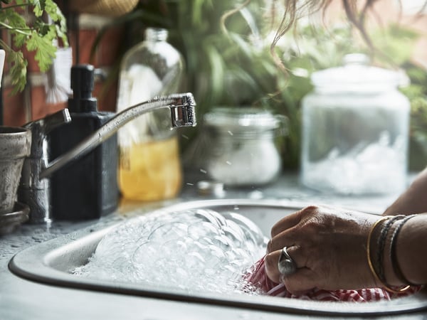 Person som tvättar grönsaker i en vask i rostfritt stål under en rinnande kran. Diskhon är omgiven av växter och olika köksartiklar.