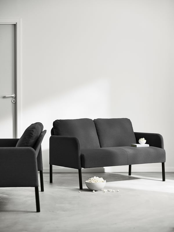 Svart modern soffa med armstöd och ryggkuddar står mot en slät vägg. Liten skål och spillde popcorn på golvet bredvid.