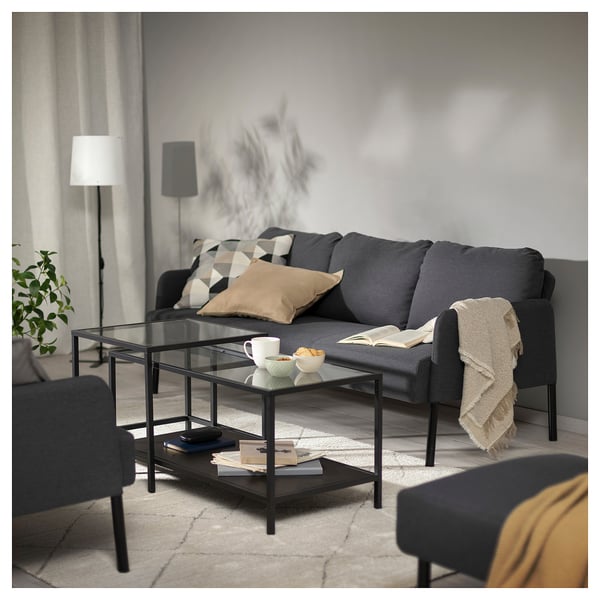 Modern grå GLOSTAD soffa med kuddar, bord med glasskiva, kaffebänk och golvlampa i minimalistiskt vardagsrum.
