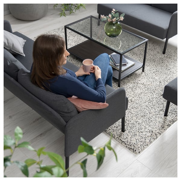 En kvinna som sitter på en GLOSTAD-grå soffa, dricker ur en mugg, med ett soffbord och en annan soffa i närheten i en mysig vardagsrumsmiljö.