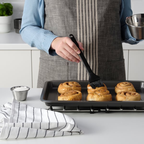 En person borstar bakverk med en silikonborste. En bakplåt och ingredienser står på bordet.