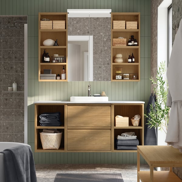 Modernt badrum med ljust hagånskåp i trä ovanför handfatet, med handdukar och flaskor.
