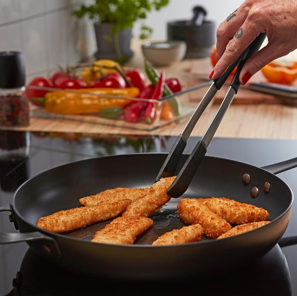 Person som lagar panerad fisk i en svart stekpanna, som håller en tång, omgiven av färgglada grönsaker.