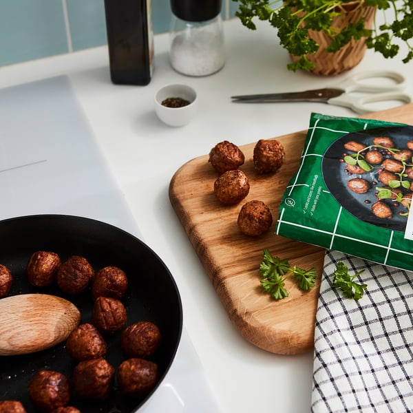 En matlagningsscen med köttbullar i en svart stekpanna och på en skärbräda av trä. Det finns en receptbok, köksredskap och en grön växt.