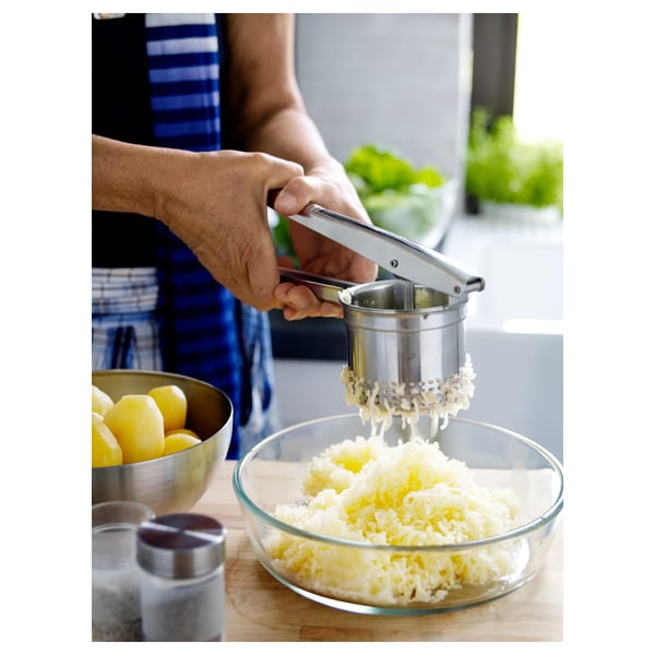 Person som använder potatispress i rostfritt stål för att mosa kokt potatis i skålen. Köksbänk med citroner, salt och örter synliga.