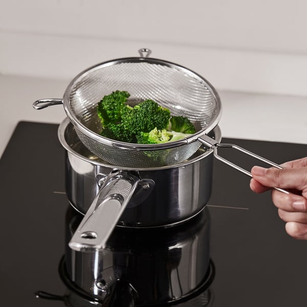 En hand håller en rostfri stålsil fylld med broccoli över en kastrull på spisen och visar hur den används för att dränera grönsaker.