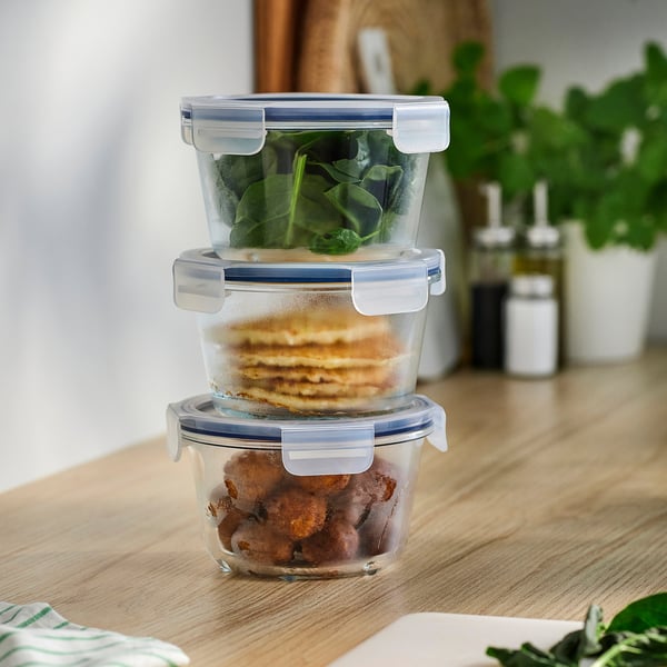 Tre staplade IKEA 365+ glasbehållare, genomskinliga, med blå lock. Rymmer mat, passar snyggt för förvaring. På träbord med växter.