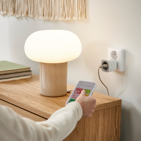 En person håller i en smartphone med en smart plug-app och styr en beige lampa på ett träbord.