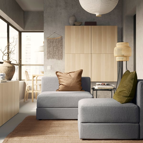 Modernt vardagsrum med grå JÄTTEBO modulsoffa, bruna prydnadskuddar, träskåp och golvlampa. Minimalistisk inredning med naturligt ljus.
