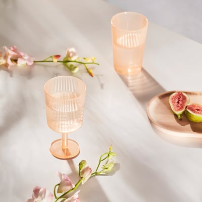 Två rosa vinglas av plast, en jordgubbe på en träplatta, omgiven av blommor på ett marmorbord.