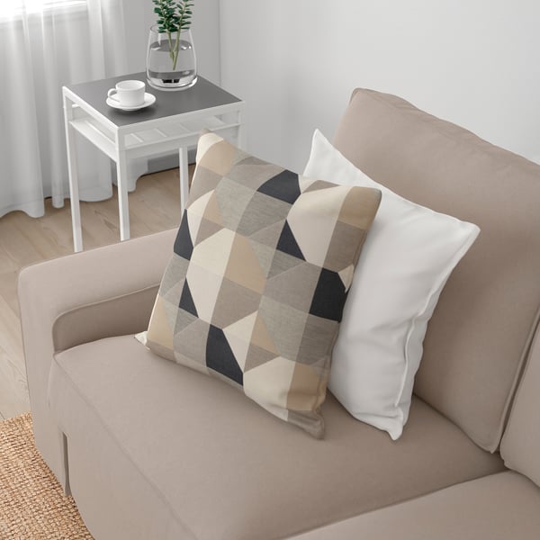 Beige KIVIK soffa med färgglad kudde, sidobord med kaffekopp, växt och vita gardiner.