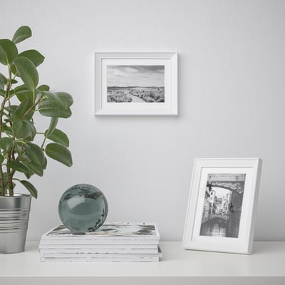 En stylad hylla rymmer en växt, en dekorativ sfär och två fotoramar, en med en kanalbild, en annan med ett landskap, mot en ljus vägg.
