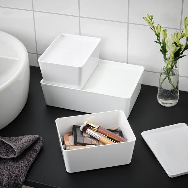 Svart disk med vita lådor, varav en med smink. Blomvas, grå handduk och handfat i närheten.