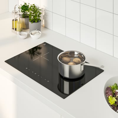Liten svart induktionsspis med kastrull, omgiven av köksartiklar, inklusive skålar, flaskor och salladsskål.