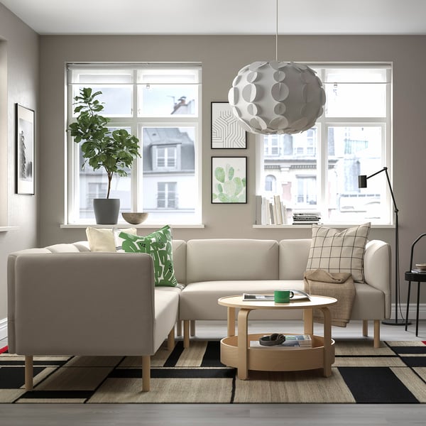 Modernt vardagsrum med en lillem l-formad beige soffa, soffbord i trä, stort fönster, hängande ljus, växt och matta.