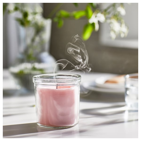 Ett brinnande rosa doftljus i en klar glasburk på ett ljust bord. Mjuk rök ringlar sig uppåt, omgiven av gröna växter och naturligt ljus.