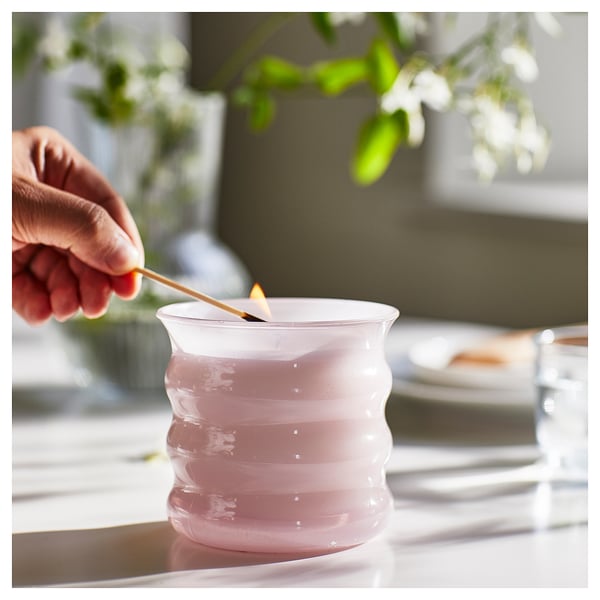 En hand rör om i ett rosa doftljus från LUGNARE i en klar glashållare på ett bord.