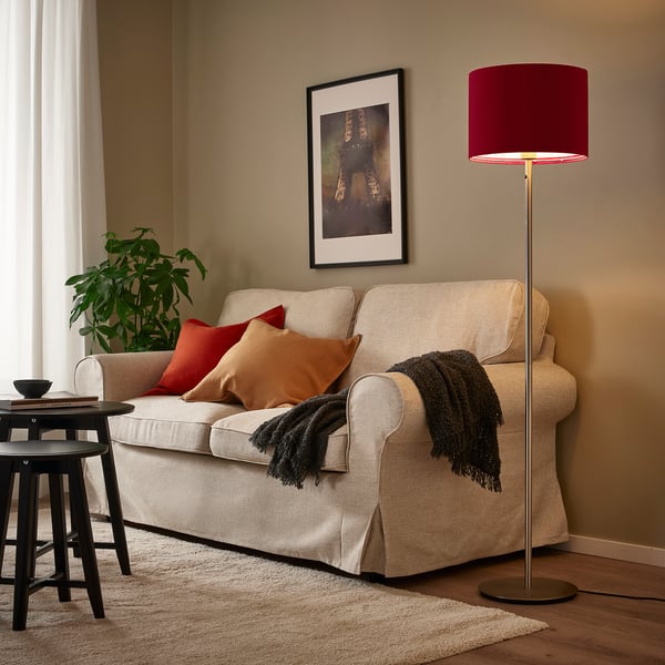 Mysigt vardagsrum med krämfärgad soffa och röda kuddar. Hög lampa med röd sammetsskärm ger värme.