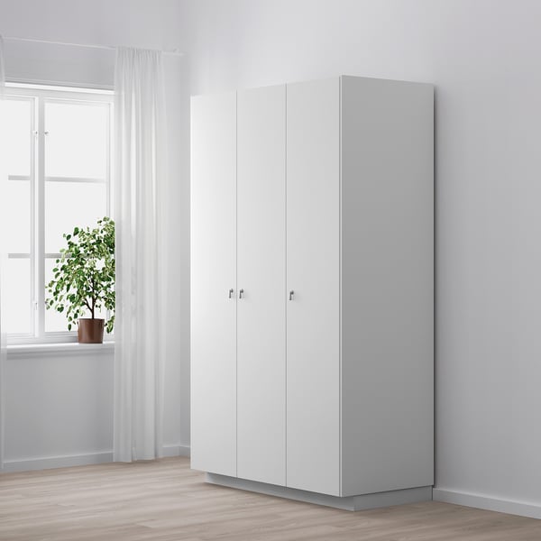 Minimalistiskt vitt skåp med silverfärgade dropphandtag, placerat nära ett fönster med skira gardiner. En krukväxt står på fönsterbrädan.